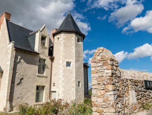 Photographie de la façade en pierre de tuffeau et schiste d'un logis ancien du XVe siècle à Blaison-Gohier. La demeure canoniale présente une tour d'escalier polygonale, des fenêtres à meneaux et une porte cochère gothique. Le bâtiment est entouré d'un jardin de curé verdoyant, avec des roses en fleurs et des vignes grimpantes, sous un ciel bleu clair. L'architecture de style Renaissance angevine est visible sur les détails des corniches et des lucarnes.