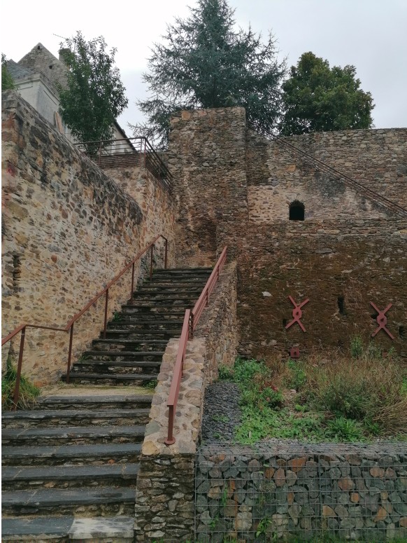 Photographie en contre-plongée des anciens remparts de schiste ardoisier de la Petite Cité de Caractère de Denée en Anjou, surmontés de maisons en tuffeau blanc, sous un ciel bleu parsemé de nuages légers.