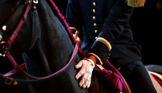 Photographie d'un écuyer du Cadre Noir de Saumur en tenue traditionnelle noire avec des dorures, montant un cheval de selle dans un manège couvert, illustrant un air de haute école (comme une courbette ou un cabré).